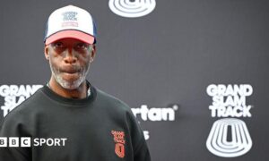 Michael Johnson looking pensive as he stands in front of a black board bearing the Grand Slam Track name and logo