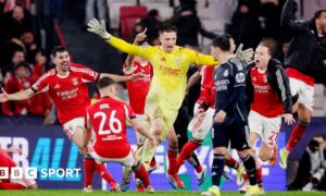 Goalkeeper Anatoliy Trubin celebrates scoring for Benfica against Real Madrid. In a yellow kit he runs among joyous team-mates who are wearing red and white kit