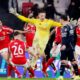 Goalkeeper Anatoliy Trubin celebrates scoring for Benfica against Real Madrid. In a yellow kit he runs among joyous team-mates who are wearing red and white kit