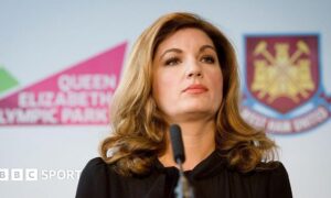 Karren Brady, with long brown hair down to her shoulders and wearing a black dress, standing in front of a microphone