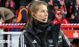 Marie-Louise Eta, wearing a black coat and top with a black microphone and headset in her right ear, on the touchline with fans in red and dark clothing in the background