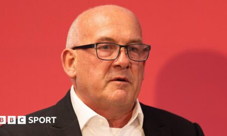 A close-up of Richard Collier-Keywood in dark jacket and open white shirt in front of a red background