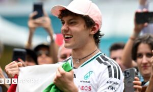 Kimi Antonelli celebrates his race win in Miami while holding the Italian flag. A crowd are gathered around him and taking photographs