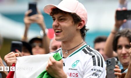 Kimi Antonelli celebrates his race win in Miami while holding the Italian flag. A crowd are gathered around him and taking photographs