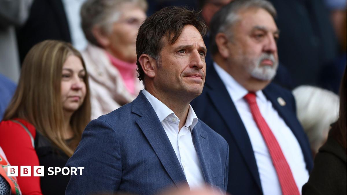 FAW chief executive Noel Mooney, in a blue suit and open neck white shirt, stands during a game.