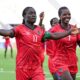 Malawi striker Tabitha Chawinga runs towards the touchline with arms spread wide to either side of her body to celebrate a goal at the 2026 Women's Africa Cup of Nations in Morocco. She wears all-red kit with green and black trim and is surrounded by a number of smiling team-mates who are running in her wake across the lush green pitch